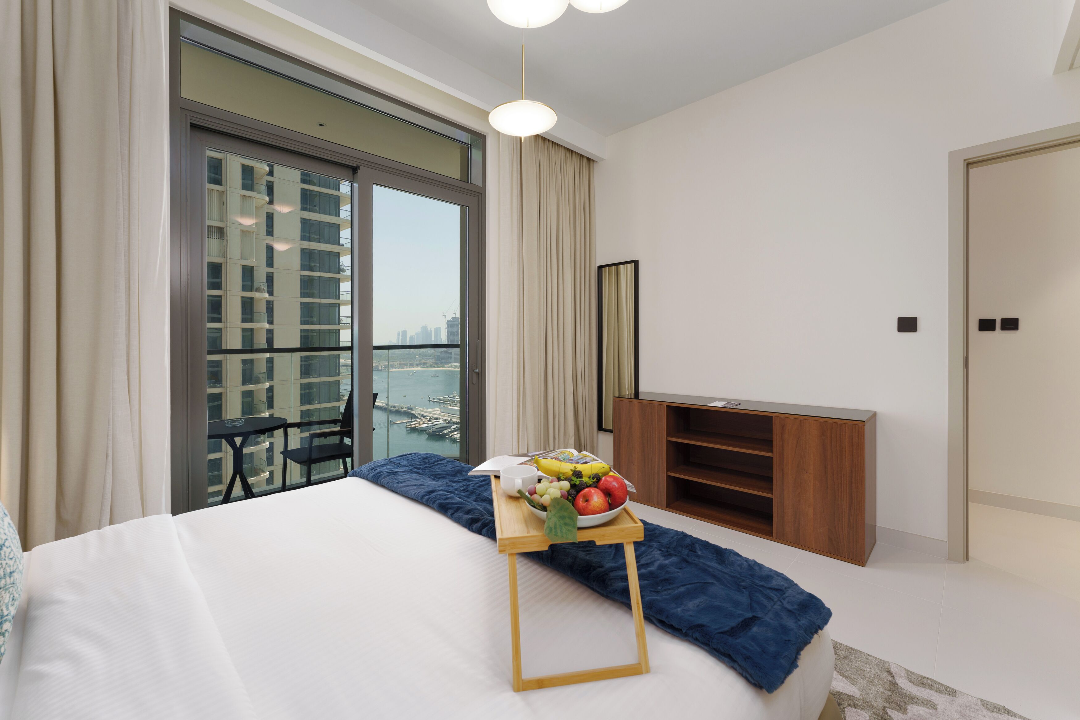 A modern hotel room with a large bed, a balcony with a city view, a wooden nightstand, and a fruit tray on the bed.
