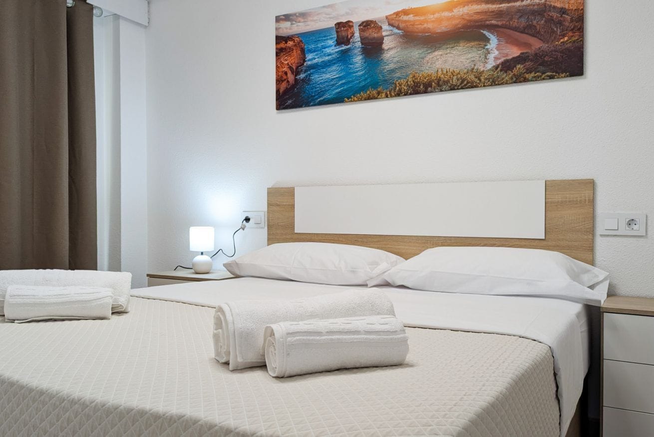 Un lit bien fait, avec du linge de lit blanc et deux serviettes enroulées, une lampe de chevet et un tableau représentant un paysage côtier accroché au mur.