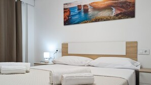 Un lit bien fait, avec du linge de lit blanc et deux serviettes enroulées, une lampe de chevet et un tableau représentant un paysage côtier accroché au mur.