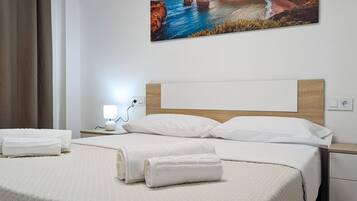 Un lit bien fait, avec du linge de lit blanc et deux serviettes enroulées, une lampe de chevet et un tableau représentant un paysage côtier accroché au mur.