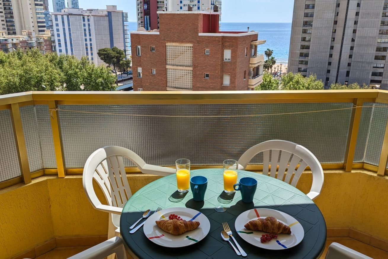 Un balcon avec deux tables, chacune portant des assiettes de croissants et des verres de jus d’orange, d’où l’on peut admirer un paysage urbain avec une plage et de hauts immeubles.