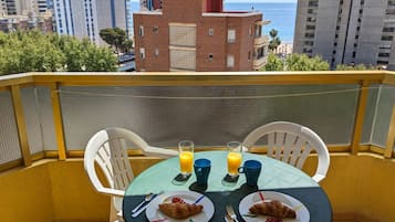 Un balcon avec deux tables, chacune portant des assiettes de croissants et des verres de jus d’orange, d’où l’on peut admirer un paysage urbain avec une plage et de hauts immeubles.