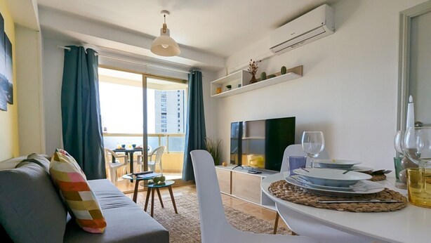 Un salon moderne avec un canapé, une table à manger pour deux personnes, un téléviseur et un balcon offrant une vue sur les immeubles.