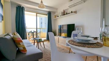 Un salon moderne avec un canapé, une table à manger pour deux personnes, un téléviseur et un balcon offrant une vue sur les immeubles.