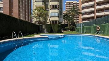Une piscine dont le fond est recouvert de carreaux disposés selon un motif géométrique, entourée d’une haie et bordée d’un immeuble imposant doté de nombreux balcons.