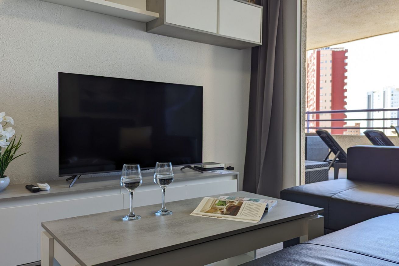A modern hotel room with a flat-screen TV, a small table with two wine glasses, a magazine, and a potted plant.