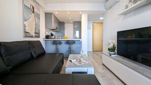 A modern living room with a black sofa, a coffee table with a magazine, a TV, and a kitchen area with bar stools. - TROPIC MAR sweet apartment (Benidorm)