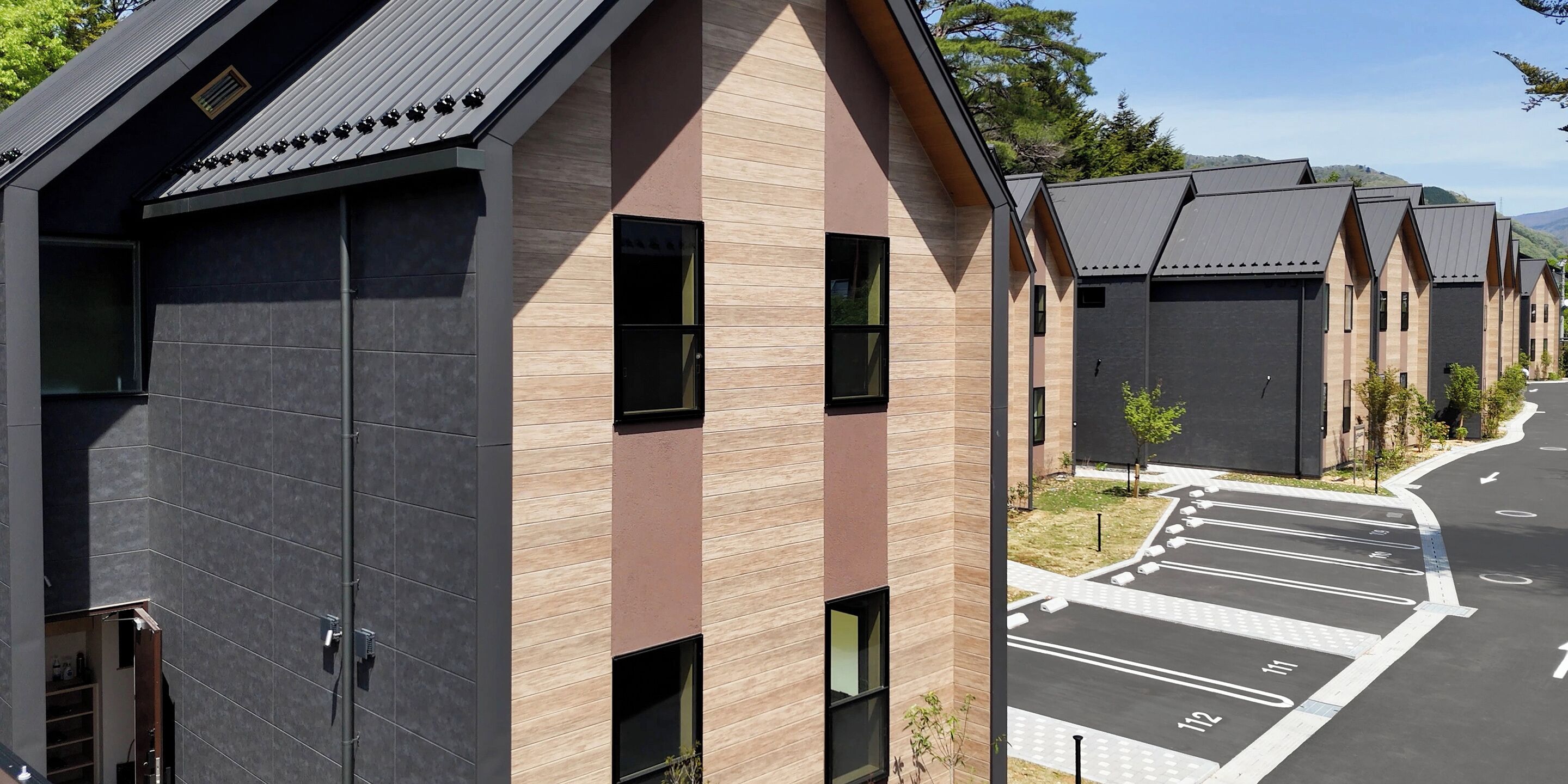 A modern building with a mix of dark and light facade panels, large windows, and a parking area with marked spaces.