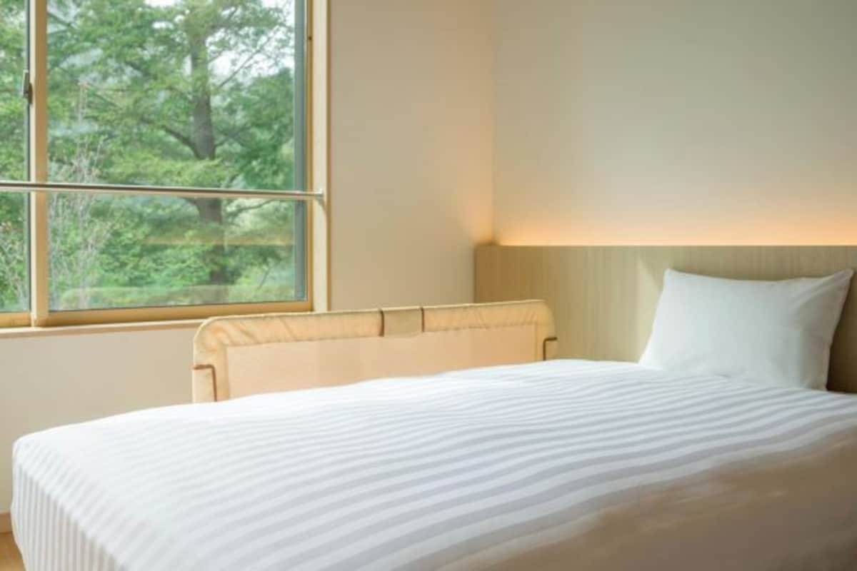 A hotel room with a bed, a window with a view of trees, and a wall-mounted lamp.
