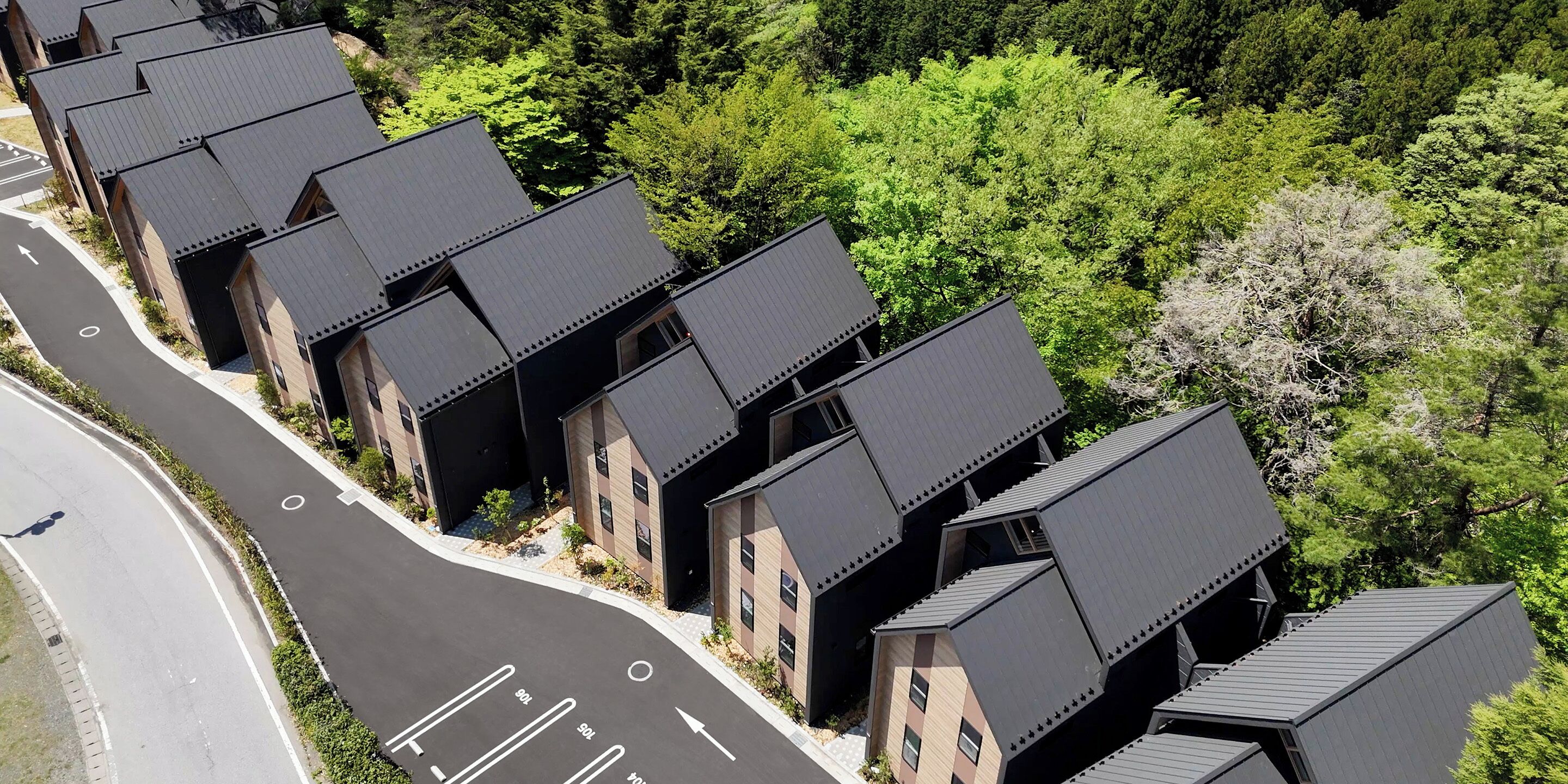 A row of modern, dark-roofed buildings with a uniform architectural style, surrounded by lush greenery and a curved road.