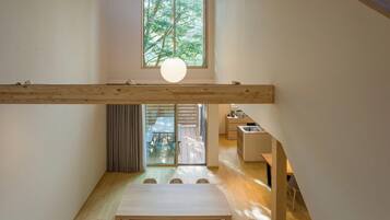 A modern interior with a high ceiling, wooden beams, a dining area with a table and chairs, and a view of a balcony with a table and chairs.