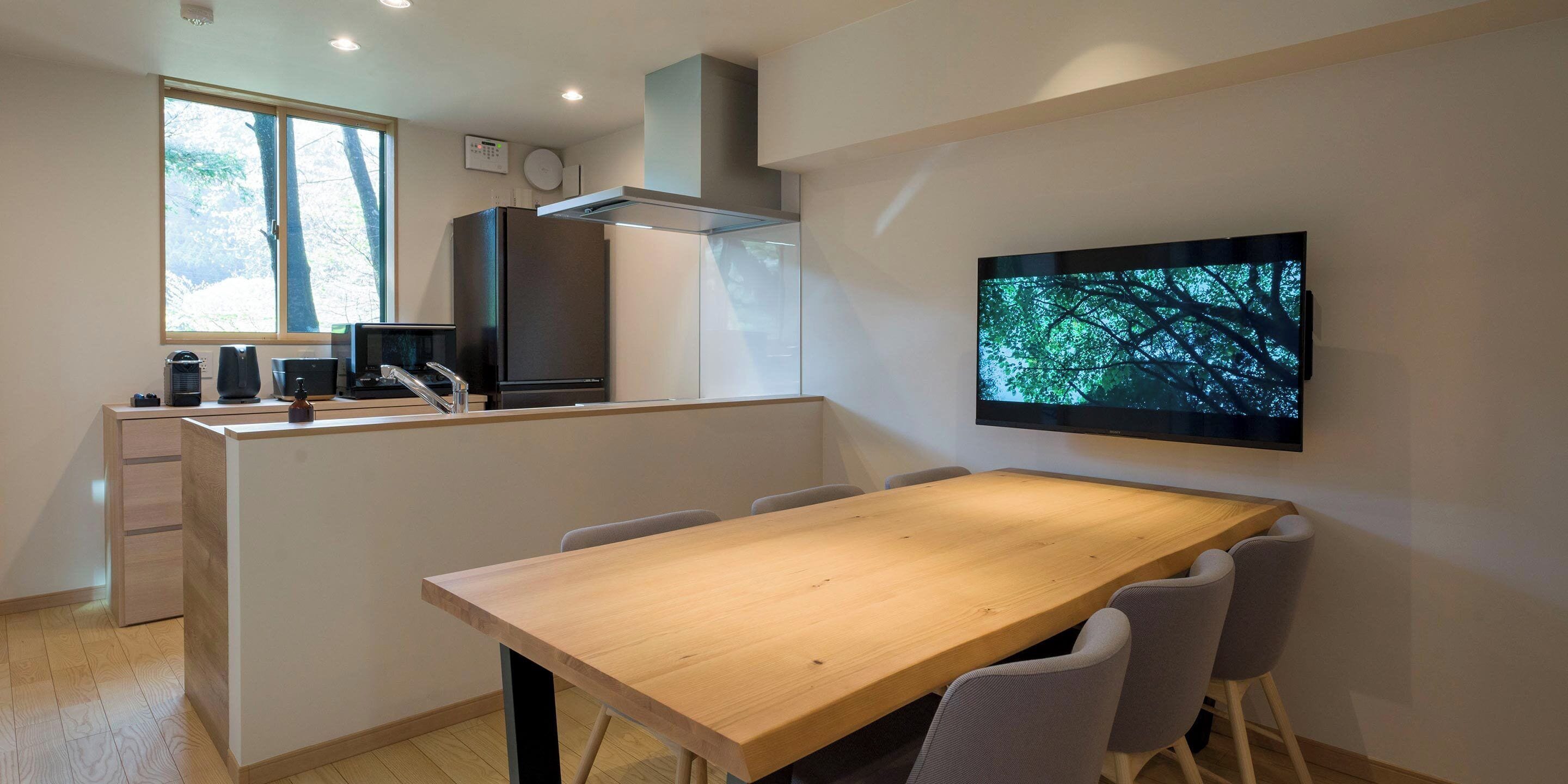 Une cuisine moderne avec une table à manger en bois, des chaises grises, un grand téléviseur fixé au mur et une fenêtre donnant sur des arbres.