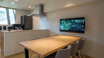Une cuisine moderne avec une table à manger en bois, des chaises grises, un grand téléviseur fixé au mur et une fenêtre donnant sur des arbres.
