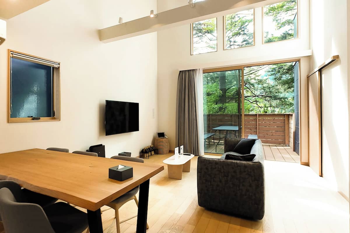 A modern interior with a wooden dining table, grey chairs, a flat-screen TV, and a large window offering a view of a garden.