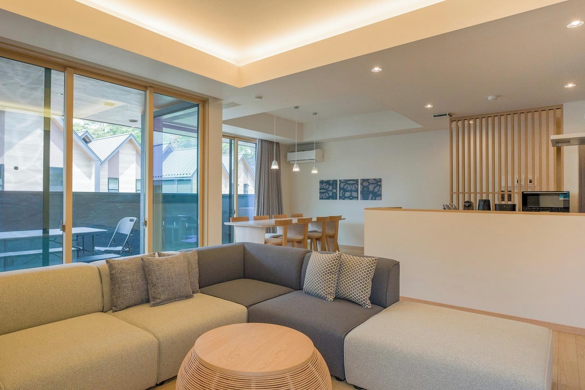 A modern living room with a sectional sofa, a round wooden coffee table, and large windows offering a view of the outside.