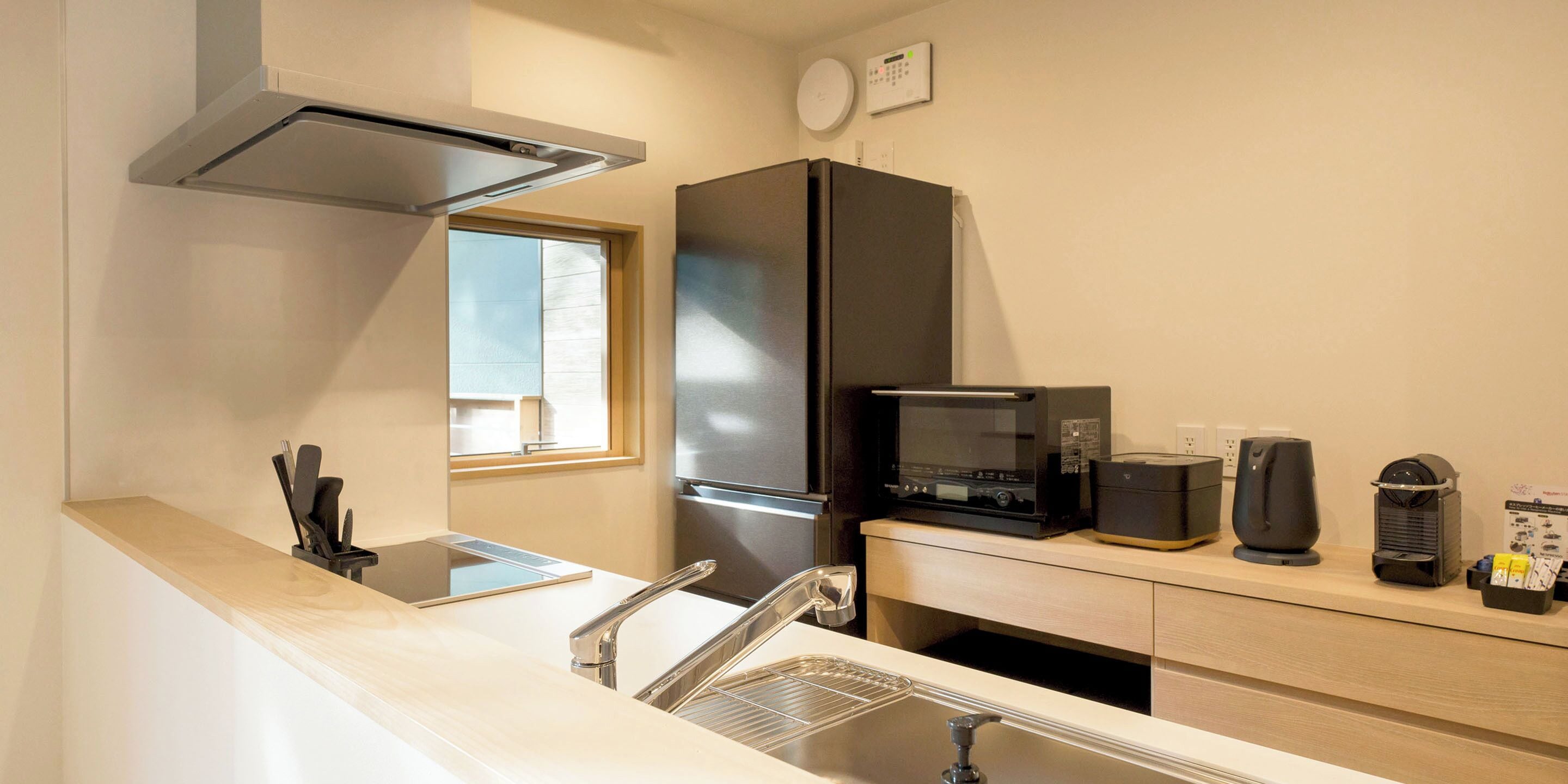 Una cocina moderna con un refrigerador de acero inoxidable, microondas y tetera. Hay una ventana que proporciona luz natural y un lavamanos con grifo.