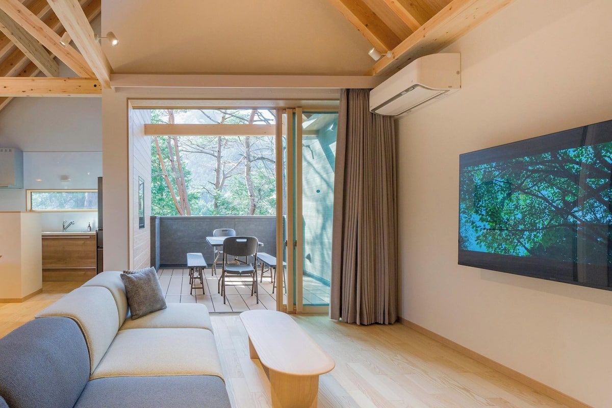 A modern living room with a sofa, dining table, and chairs. There is a kitchen area with a sink and cabinets, and a large window with curtains.