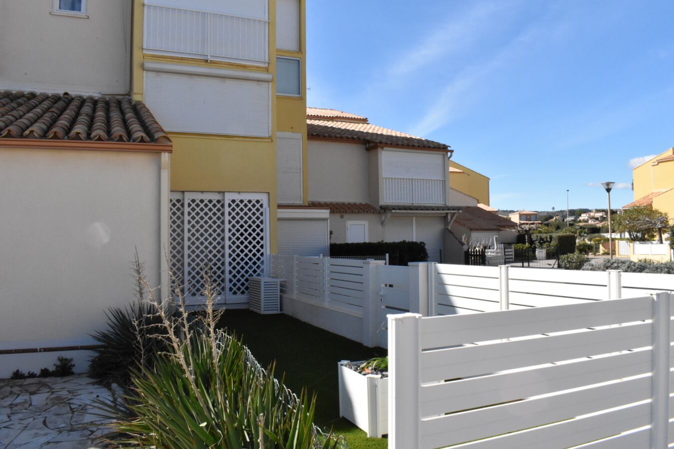A residential building with a tiled roof, a white fence, and a small garden area with plants.