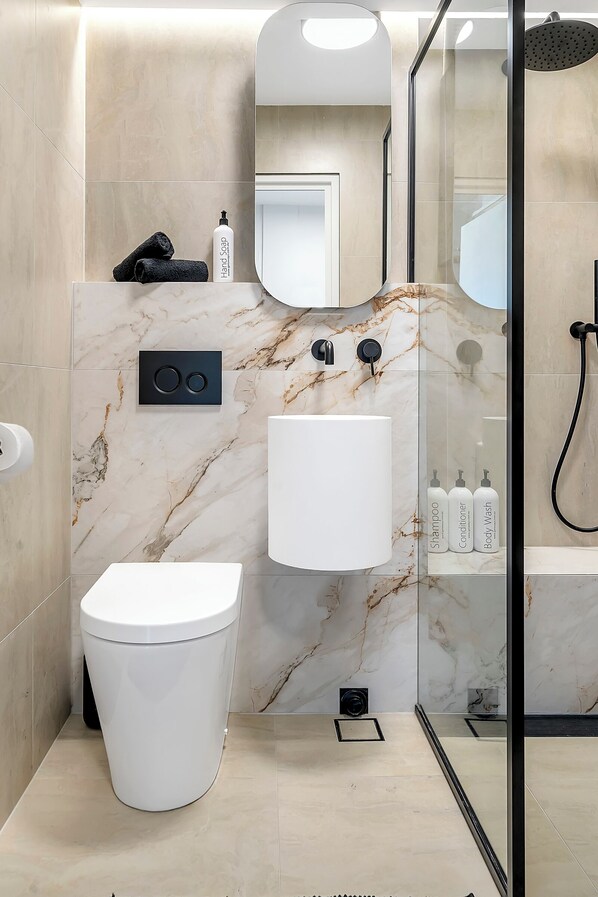 Un baño moderno con salpicadero de mármol, inodoro blanco, lavabo con espejo arriba y zona de ducha.