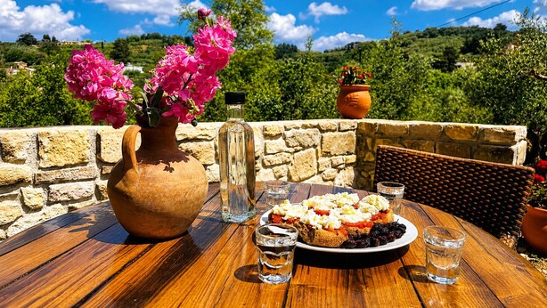 A wooden table with a plate of food, a vase with pink flowers, and a bottle. - Oniropagida - Dream Catcher - House On Way To Sougia (Platanias)