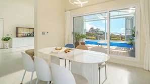 Un espace repas moderne avec une table en verre et des chaises blanches, un téléviseur fixé au mur, et une vue sur une piscine et la verdure environnante.