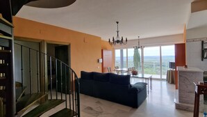 Un'ampia zona giorno con un divano blu scuro, un tavolo da pranzo e vista sulla campagna attraverso ampie finestre.