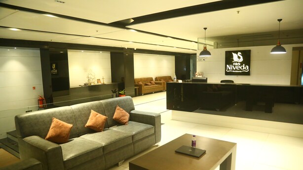 A modern hotel lobby with a grey sofa, a wooden coffee table, and a reception desk. - Park Niveda (Hyderabad)