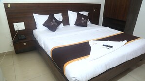 A neatly made bed with a white and brown bedspread, two white pillows, and two brown pillows with a logo. There is a bedside table with a phone and a remote control. - Park Niveda (Hyderabad)