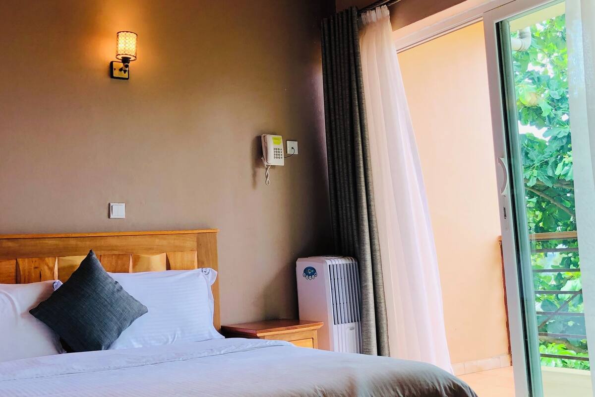 Un dormitorio con techo de madera, una cama con ropa blanca, una mesita de noche, un teléfono en la pared y una ventana con cortinas.