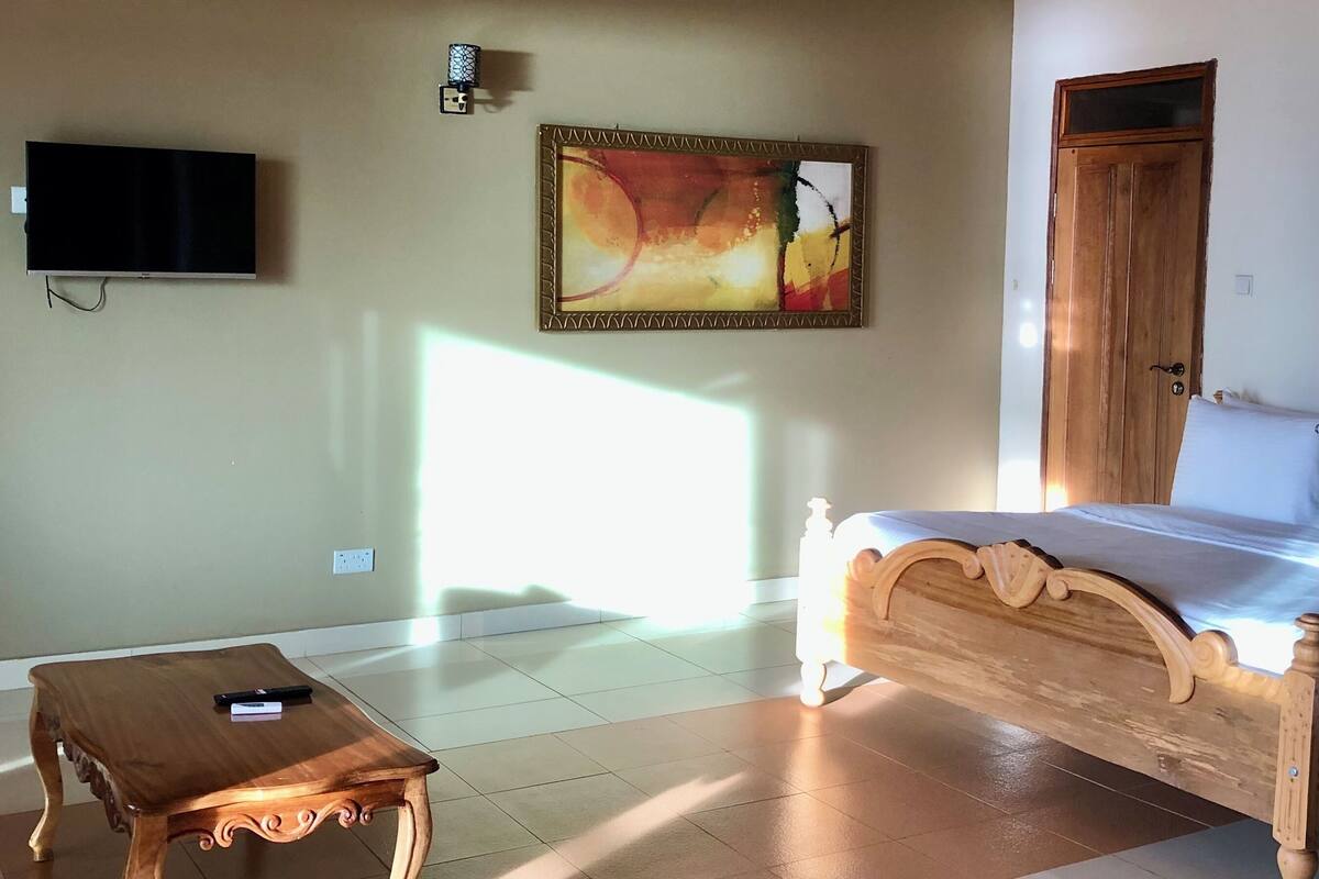 Una habitación con techo de madera, una cama con colchón blanco, una mesita de centro de madera, un cuadro en la pared y un televisor sobre la cama.