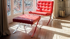 A room with a red leather chair, a wooden table, and a large window with a view of buildings. - Sun-drenched Sophistication, by Veeve (Paris)