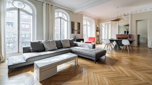 A spacious living room with a large sectional sofa, a dining area with a red chair, and a herringbone wood floor. - Sun-drenched Sophistication, by Veeve (Paris)