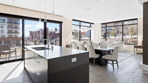 A modern kitchen with a large island, dining area with white chairs, and large windows offering views of buildings and trees.