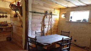 A rustic wooden interior with a wooden table and chairs, a wooden bench, a shelf with kitchen utensils, and a window with a view.