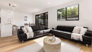 A modern living room with a sectional sofa, a round coffee table, and a large window with a view of greenery. - Lamington's Landing near Seacliff Beach (Seacliff Park)