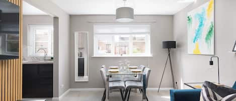 A modern dining area with a glass table, grey chairs, and a large window with blinds.