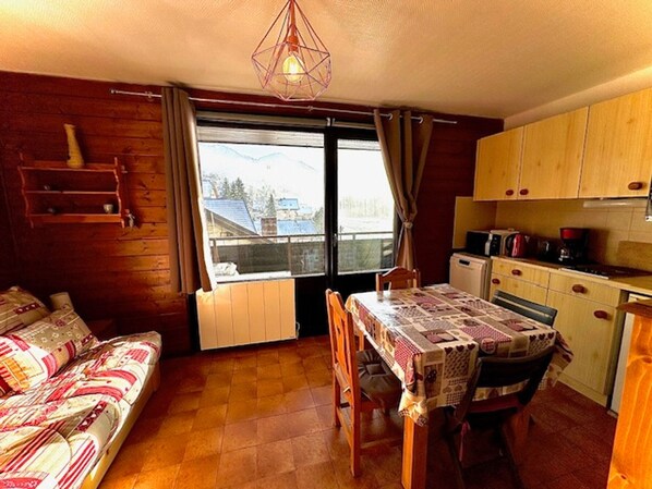 A cozy room with a wooden floor, a dining table with chairs, a sofa, a kitchen area with cabinets, and a window with curtains. - Studio in the center, near lifts! (La Chapelle-d'Abondance)
