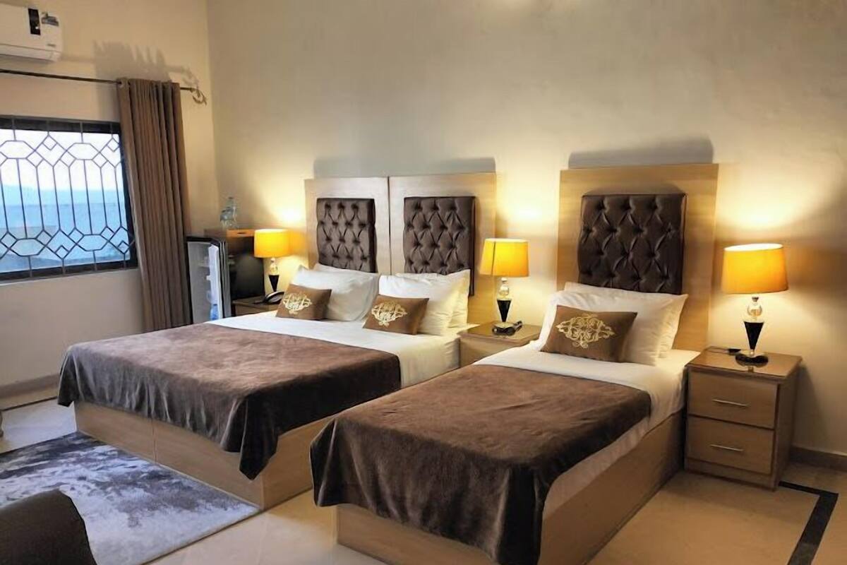 Ein Hotelzimmer mit zwei Betten, jeweils mit Kopfteil, Nachttischen mit Lampen und einem Fenster mit Vorhängen.