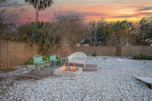 A backyard with a fire pit, two green metal chairs, a white plastic chair, and a wooden deck.
