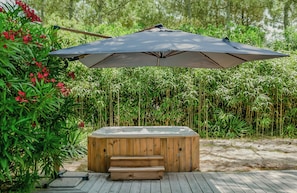 Een hottub met een houten buitenkant, omringd door weelderige begroeiing en een grote parasol voor schaduw.