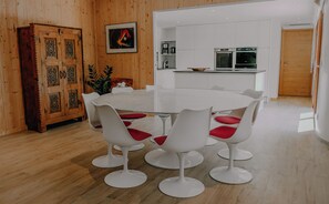 Un comedor con una mesa redonda y sillas blancas con cojines rojos, un mueble de madera con diseños intrincados y una cocina al fondo.