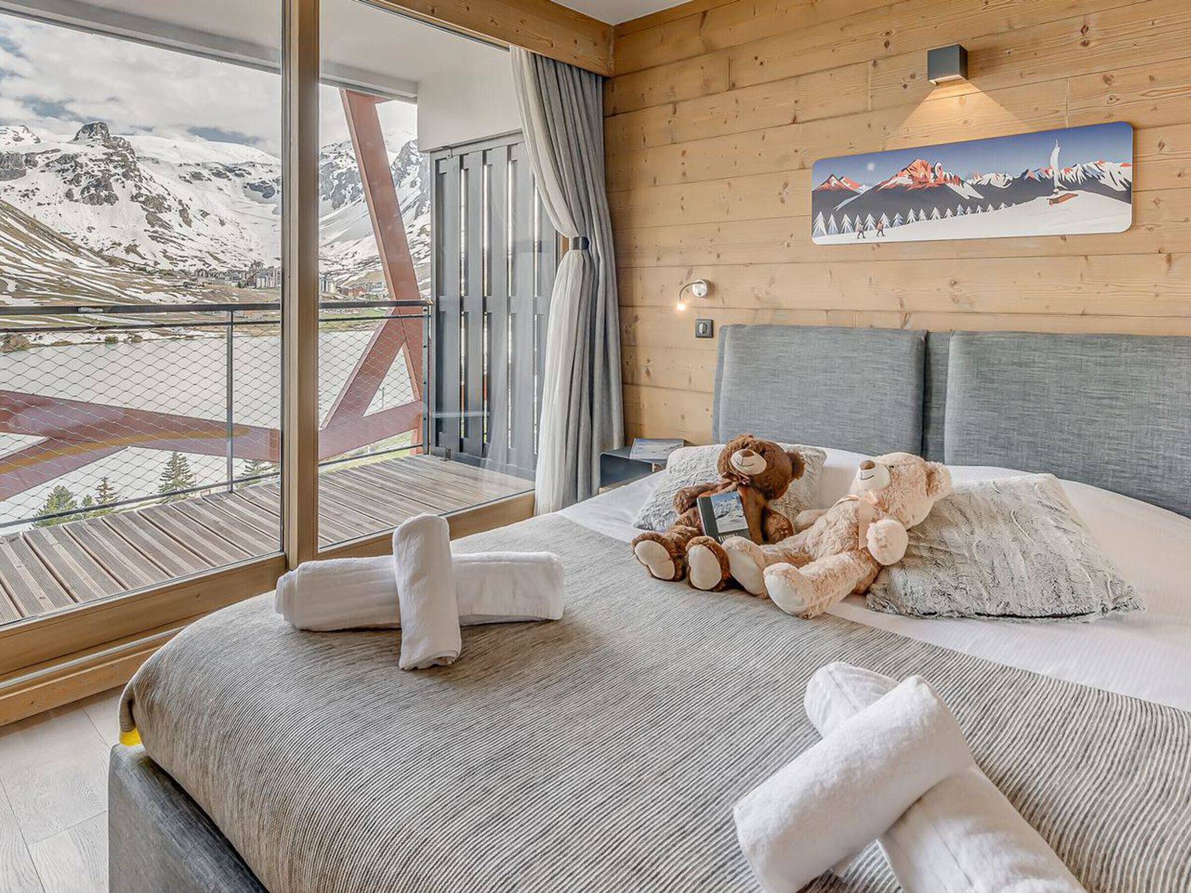 Et hotelværelse med et stort vindue med udsigt over et sneklædt bjerglandskab, en seng med to bløde bamser og et bjergmaleri på væggen.
