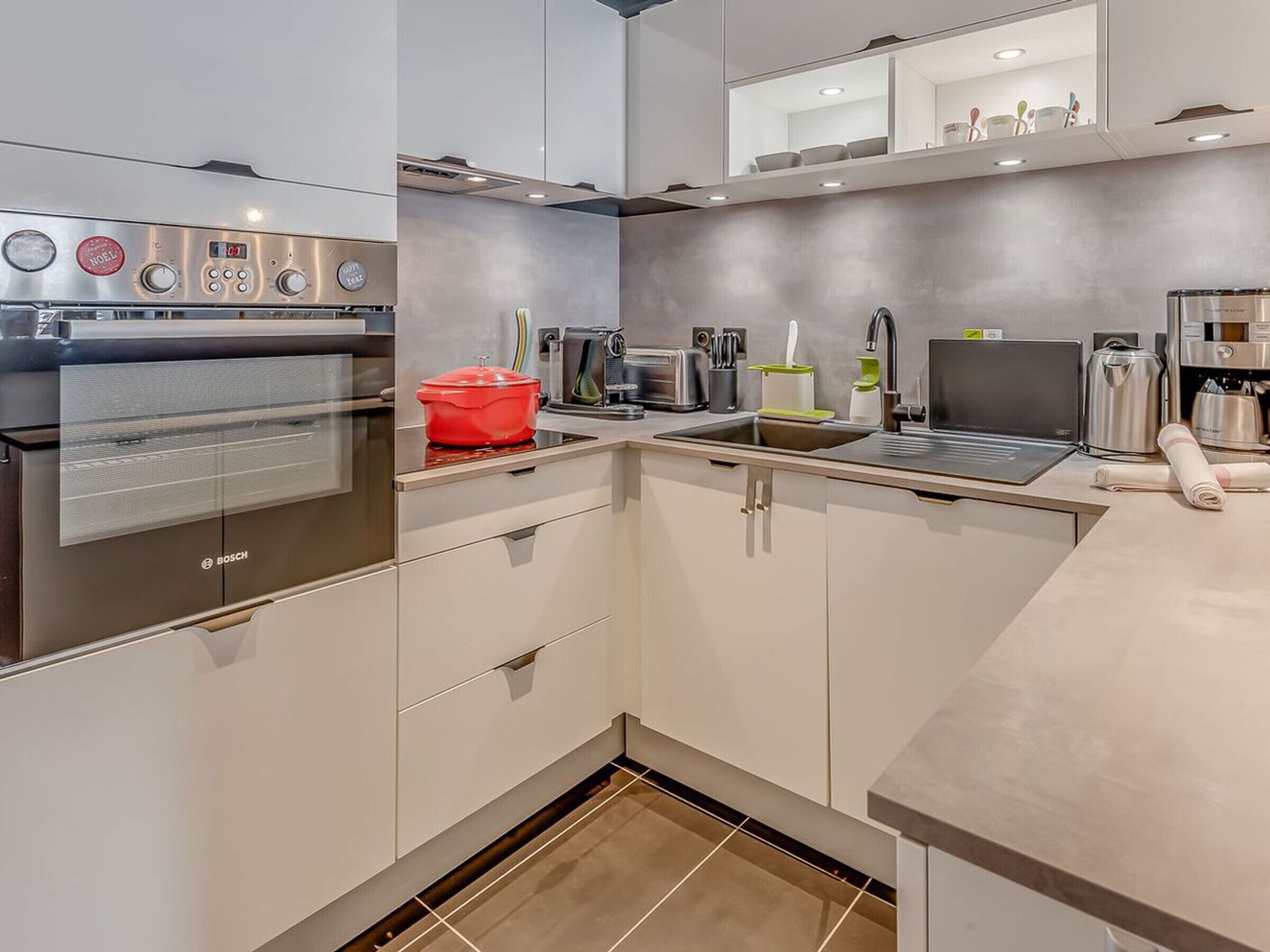 A modern kitchen with a Bosch oven, a red pot on the stove, a sink, a coffee maker, and a laptop on the counter.