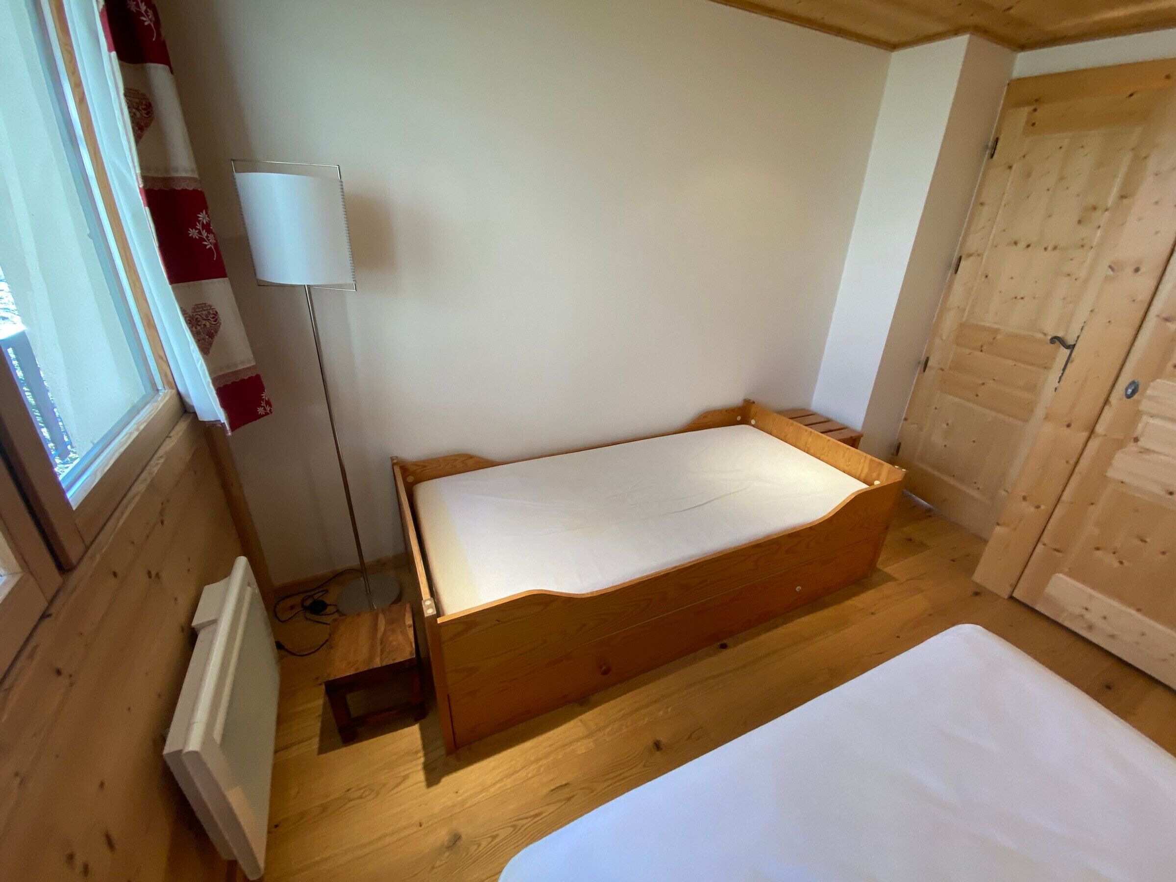 A small, wooden room with a single bed, a radiator, a window, and a door.