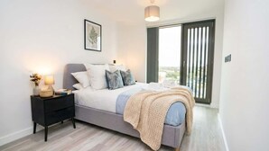 A modern bedroom with a bed, bedside table, lamp, and a view of the outdoors through a large window. - Bright & Stylish Central Birmingham 2 Bed Flat (Birmingham)