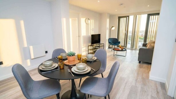 A modern dining area with a round table set for four, surrounded by grey chairs. A television is mounted on the wall, and a sofa is visible in the background. - Bright & Stylish Central Birmingham 2 Bed Flat (Birmingham)