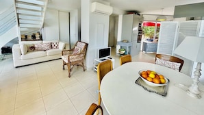 A spacious living area with a white sofa, a wicker chair, a dining table with a fruit bowl, and a staircase leading to another level.