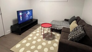 Una sala de estar con un televisor sobre un mueble bajo, una mesa roja, un tapete estampado y un sofá marrón con cojines.