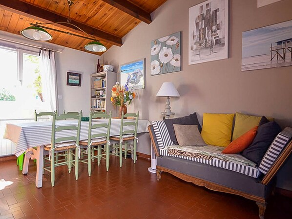 A cozy living room with a wooden ceiling, a dining area with a table and chairs, and a sofa with patterned cushions.