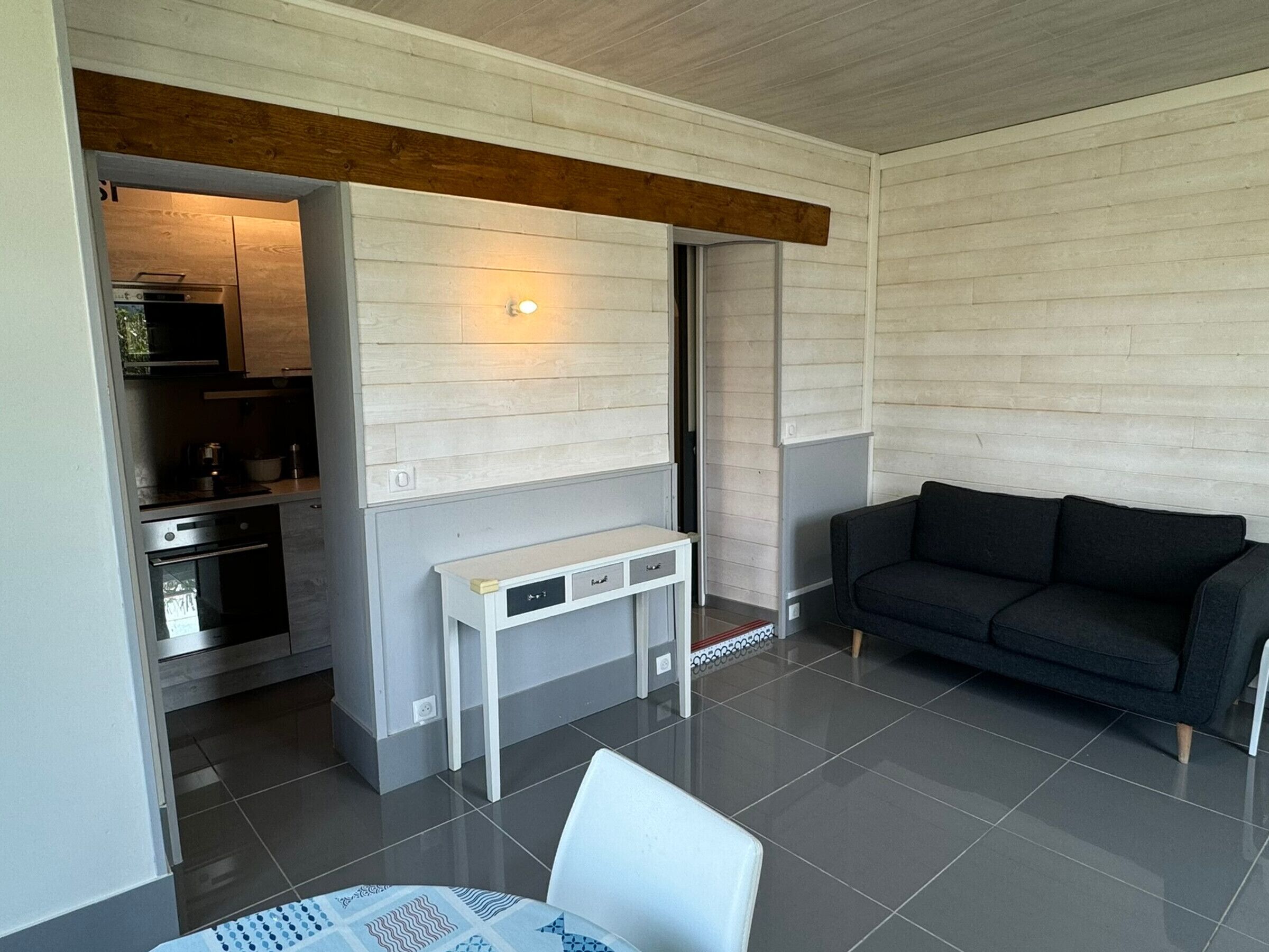 En moderne stue med en mørk sofa, et hvidt bord og et køkkenområde, der ses gennem en åben dør.